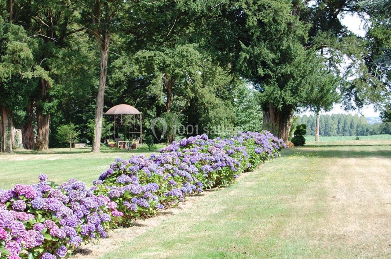 Allée de fleurs d'hortensias violets bordant une pelouse, avec un kiosque en arrière-plan sous des arbres.