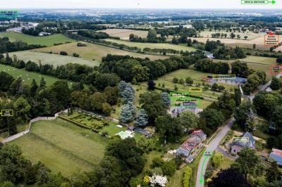 Vue aérienne d'une campagne avec des champs, des arbres, des maisons et des routes annotées pour indiquer des directions.