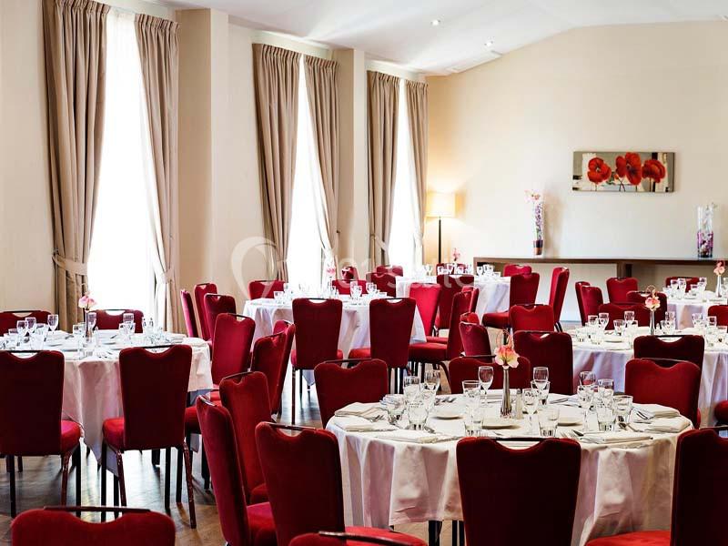 Salle de réception lumineuse avec des tables rondes dressées, entourées de chaises rouges et décorée de rideaux beiges.