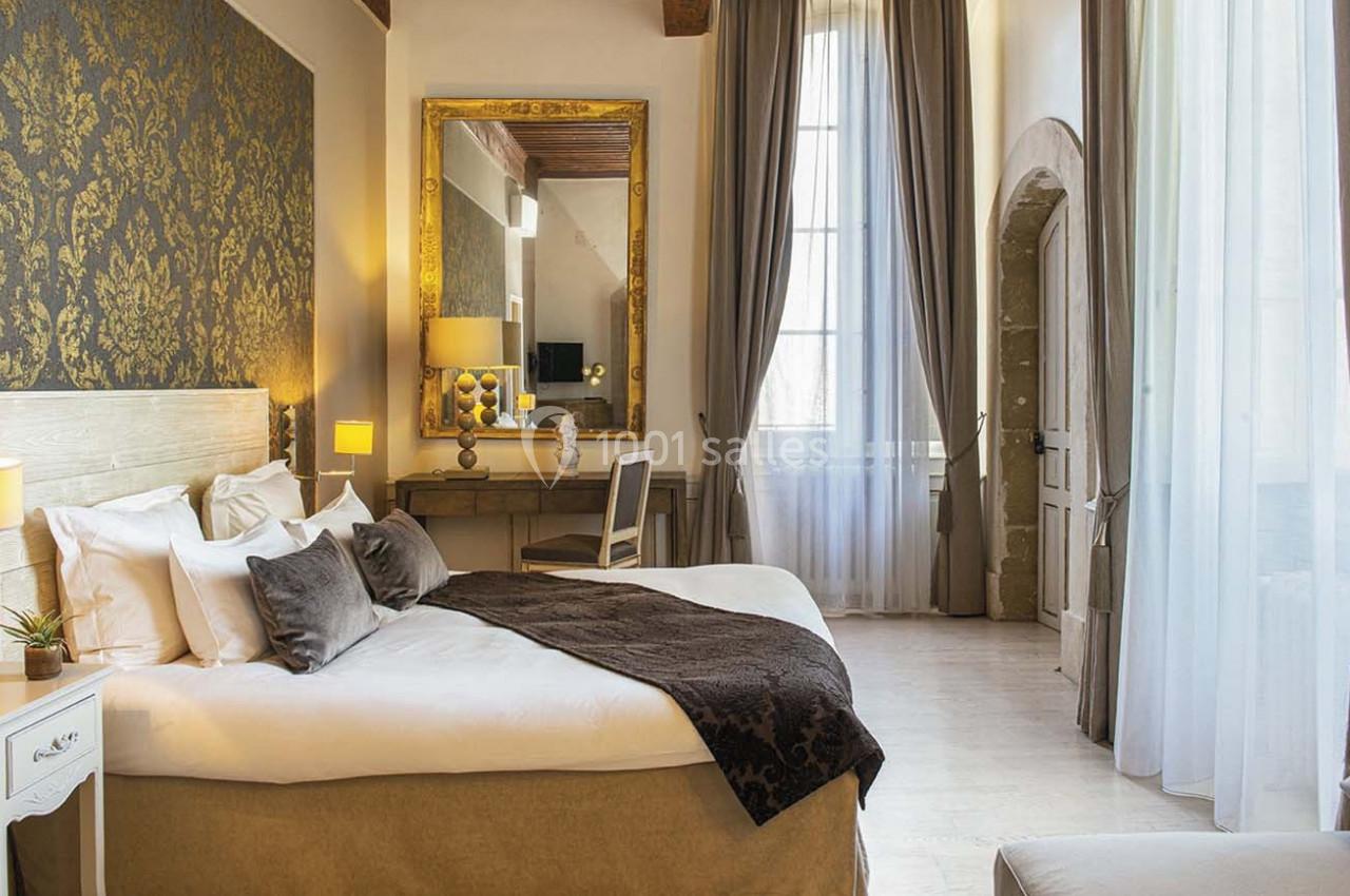 Chambre lumineuse avec lit double, décor élégant, murs ornés de motifs dorés et grande fenêtre avec rideaux.