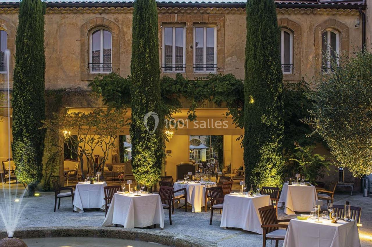 Cour intérieure d'un restaurant avec des tables dressées, entourée de cyprès et d'une façade en pierre éclairée.