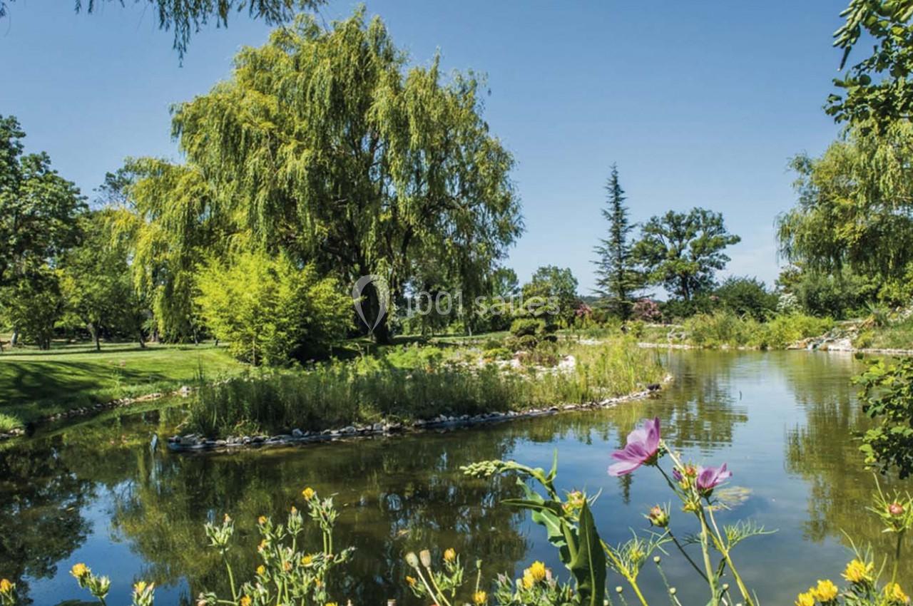 Étang entouré de verdure avec des arbres, des fleurs au premier plan et un ciel bleu dégagé.