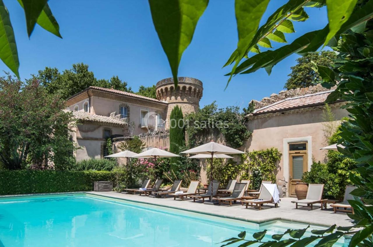 Piscine extérieure entourée de chaises longues, parasols, végétation luxuriante et bâtiments en pierre avec une tour.