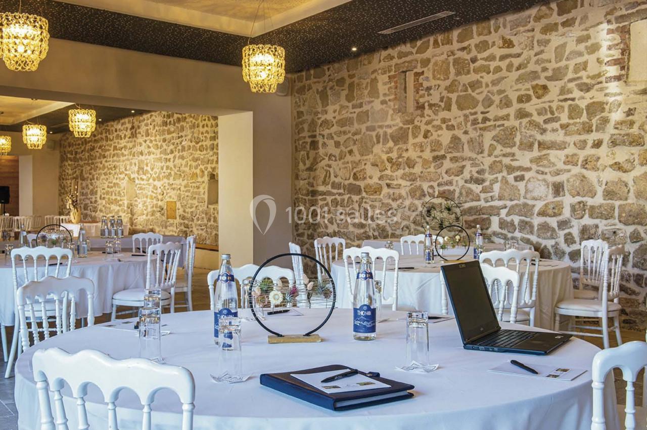 Salle de réception avec murs en pierre, tables rondes dressées et décorations lumineuses suspendues.