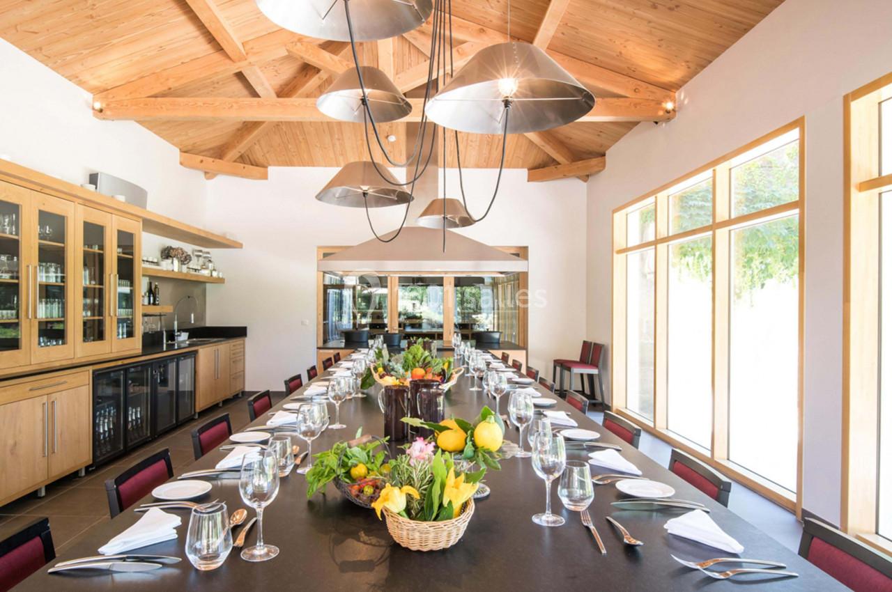 Grande table dressée avec des couverts et des paniers de fruits et fleurs, dans une salle lumineuse au plafond en bois.