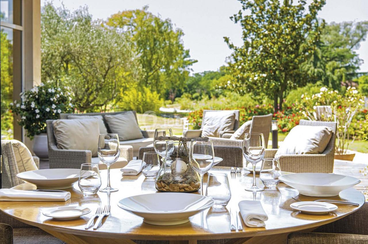 Table dressée avec vaisselle blanche et verres, donnant sur un jardin verdoyant avec mobilier d'extérieur.