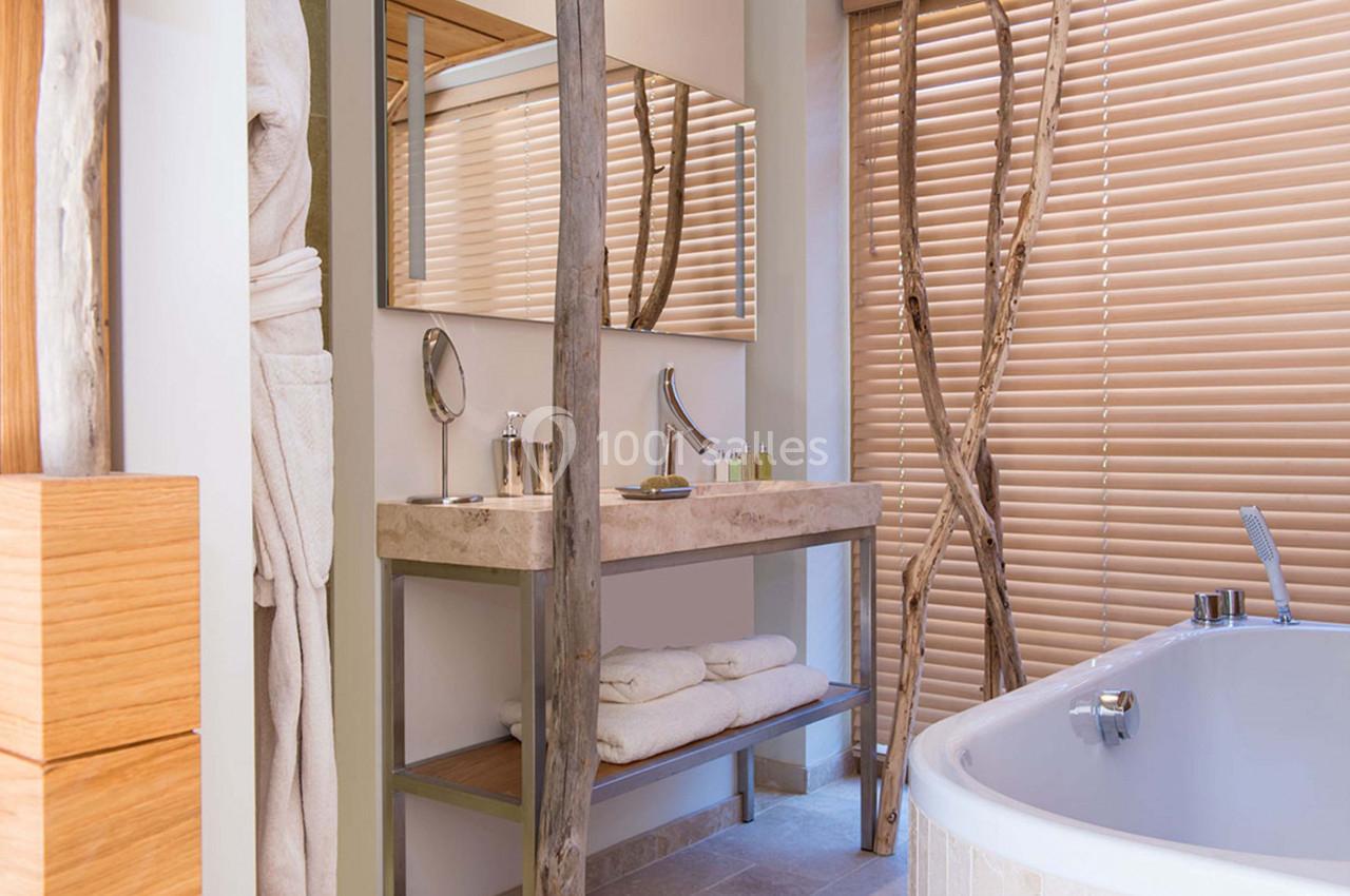 Salle de bain moderne avec lavabo en pierre, baignoire, miroir, serviettes pliées et stores en bois.