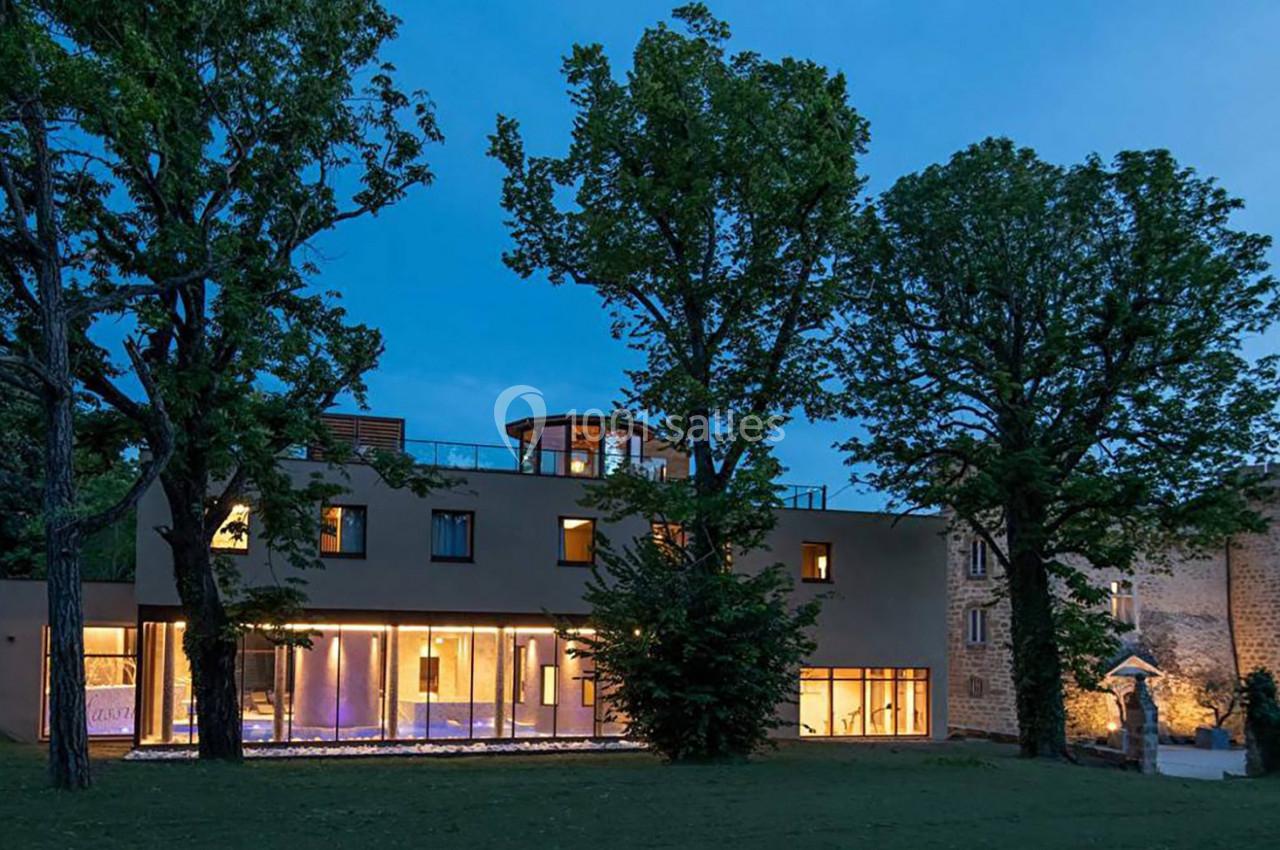 Bâtiment moderne éclairé à la tombée de la nuit, entouré d'arbres et jouxtant une structure en pierre ancienne.