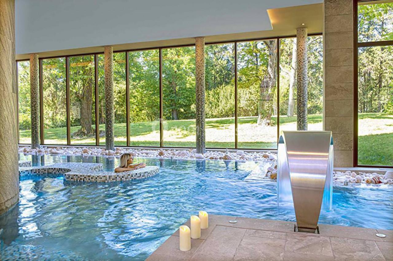 Piscine intérieure avec jets d'eau, baigneuse allongée sur une plateforme, grandes baies vitrées donnant sur un jardin.