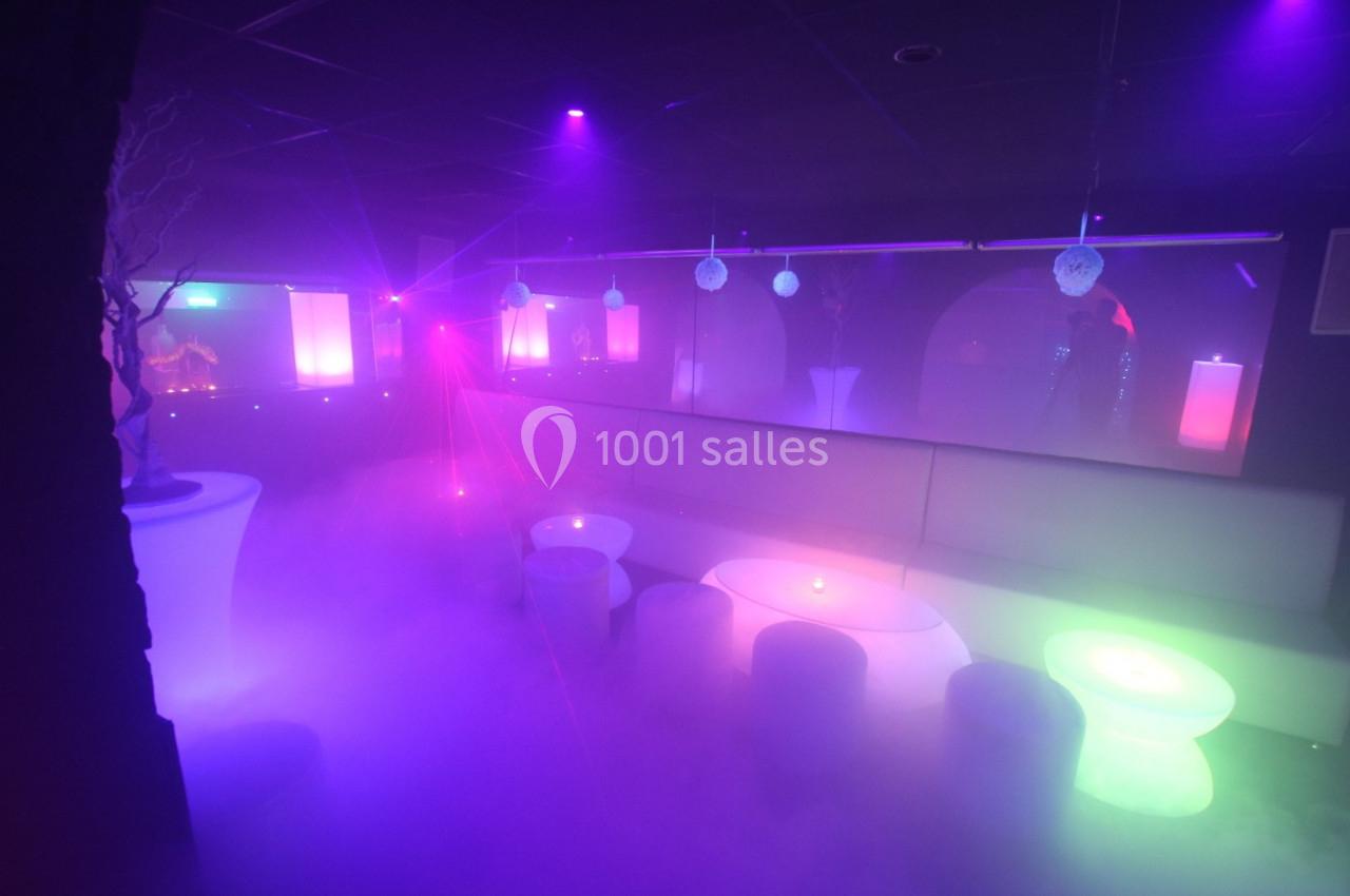 Salle de nuit avec éclairage violet et néons colorés, tables lumineuses et ambiance enfumée.