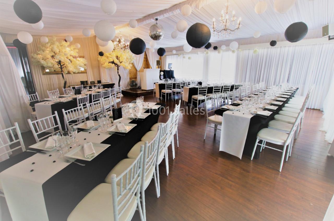 Salle de réception décorée en noir et blanc avec tables dressées, chaises blanches et lanternes suspendues.