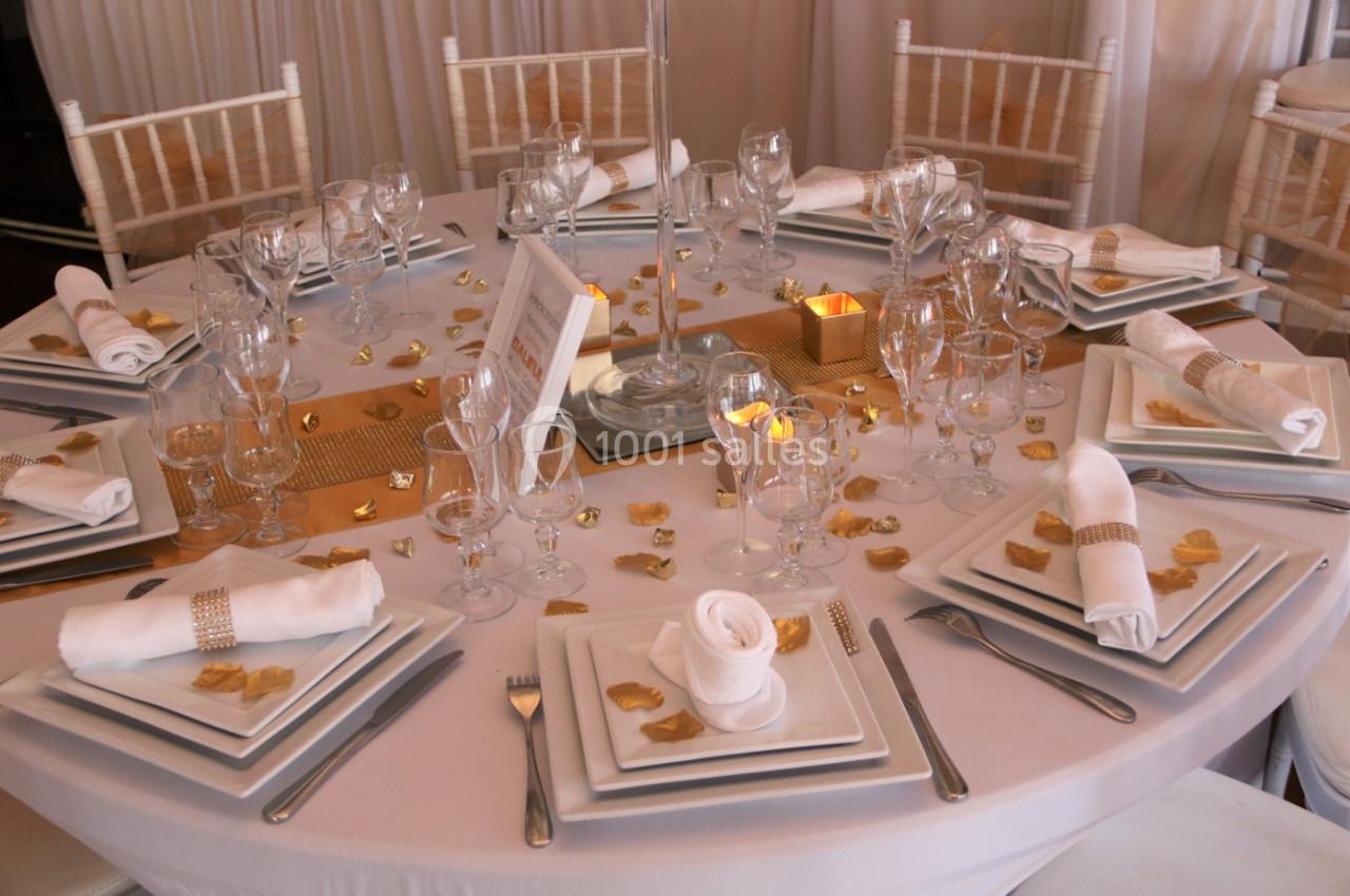 Table ronde élégamment dressée avec vaisselle blanche, serviettes pliées, décorations dorées et bougies.