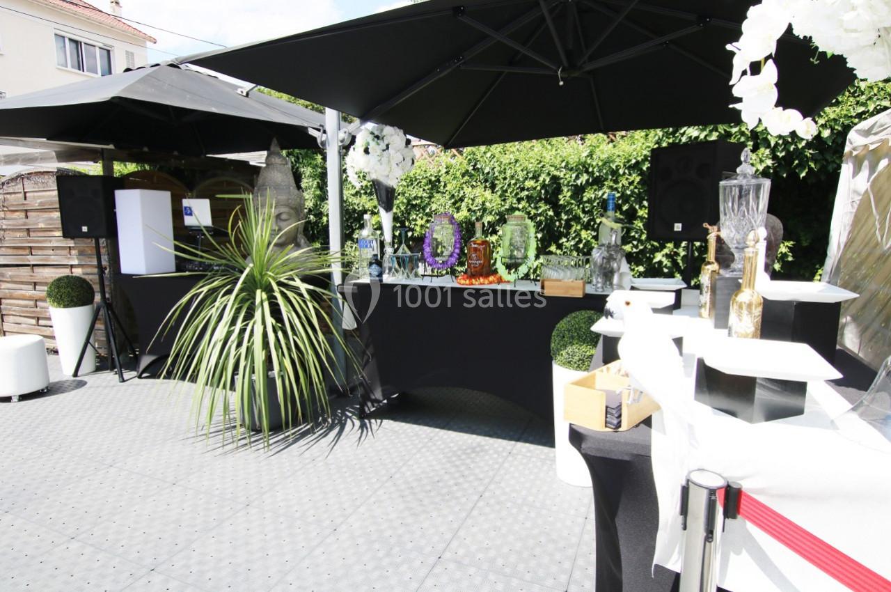 Bar extérieur avec comptoir noir, décorations florales, plantes, fontaines à boissons et parasols noirs.