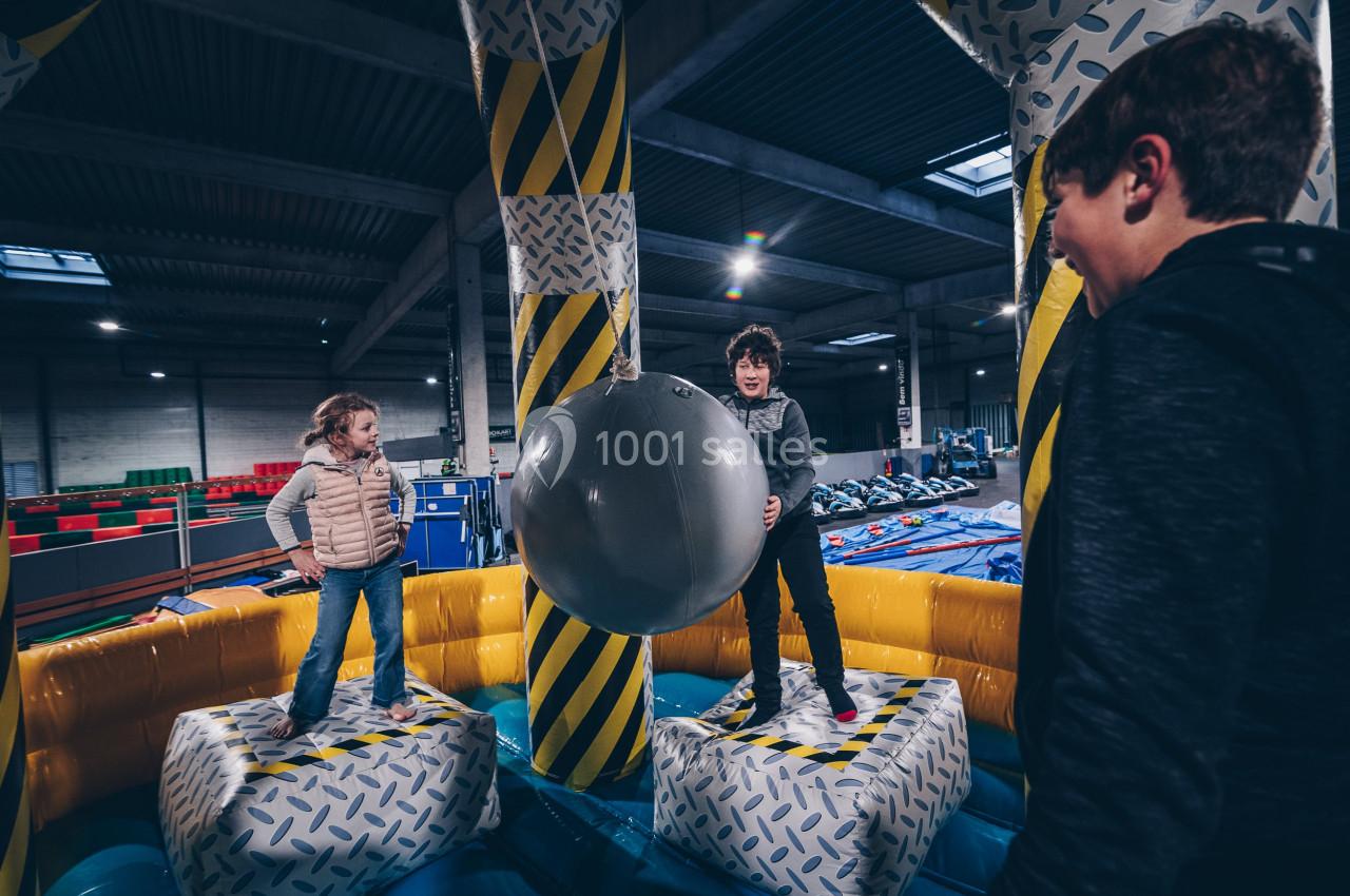 Deux enfants jouent sur une structure gonflable avec une grande balle suspendue, sous le regard d'un adulte.
