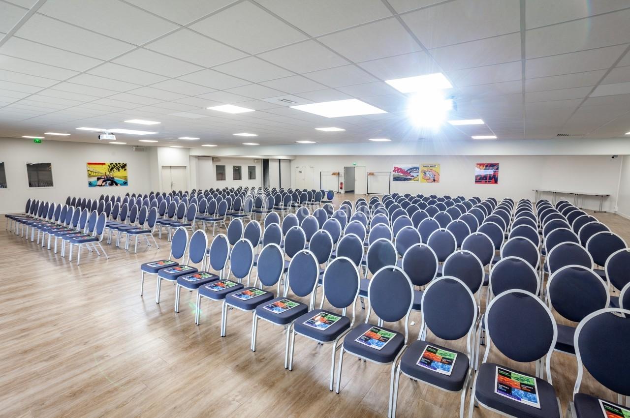 Salle de conférence lumineuse avec rangées de chaises bleues alignées et brochures posées sur certaines assises.