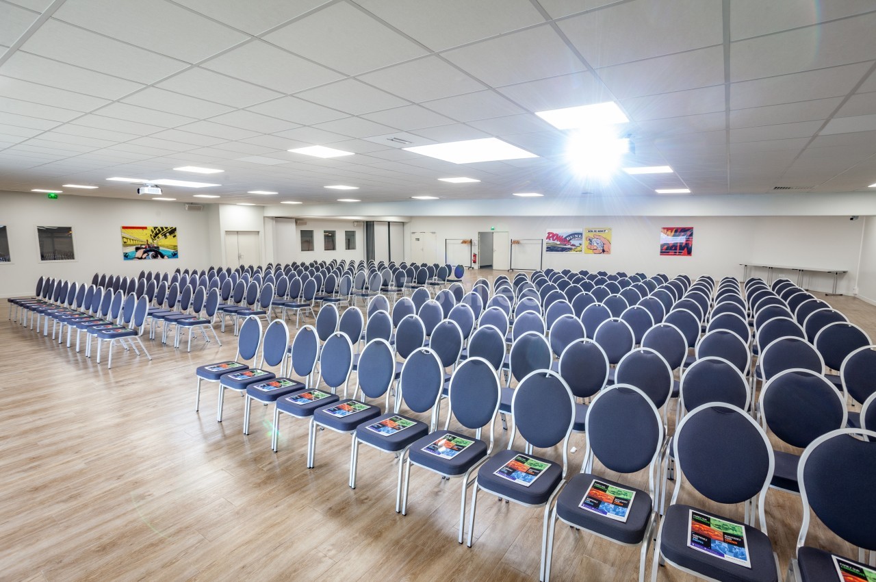 Salle de conférence lumineuse avec rangées de chaises bleues alignées et brochures posées sur certaines assises.
