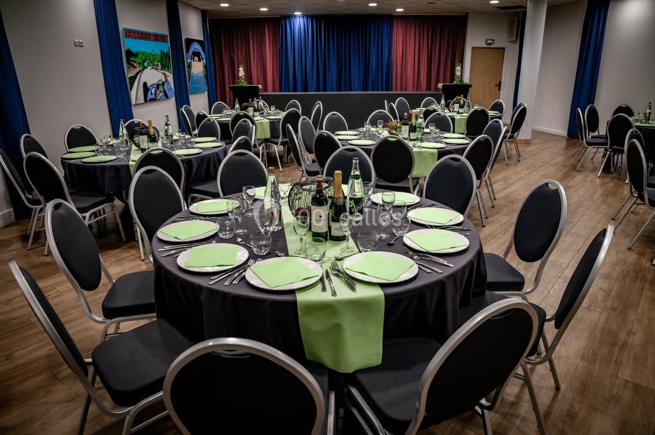 Salle de réception avec tables rondes dressées, nappes noires, serviettes vertes et bouteilles disposées.