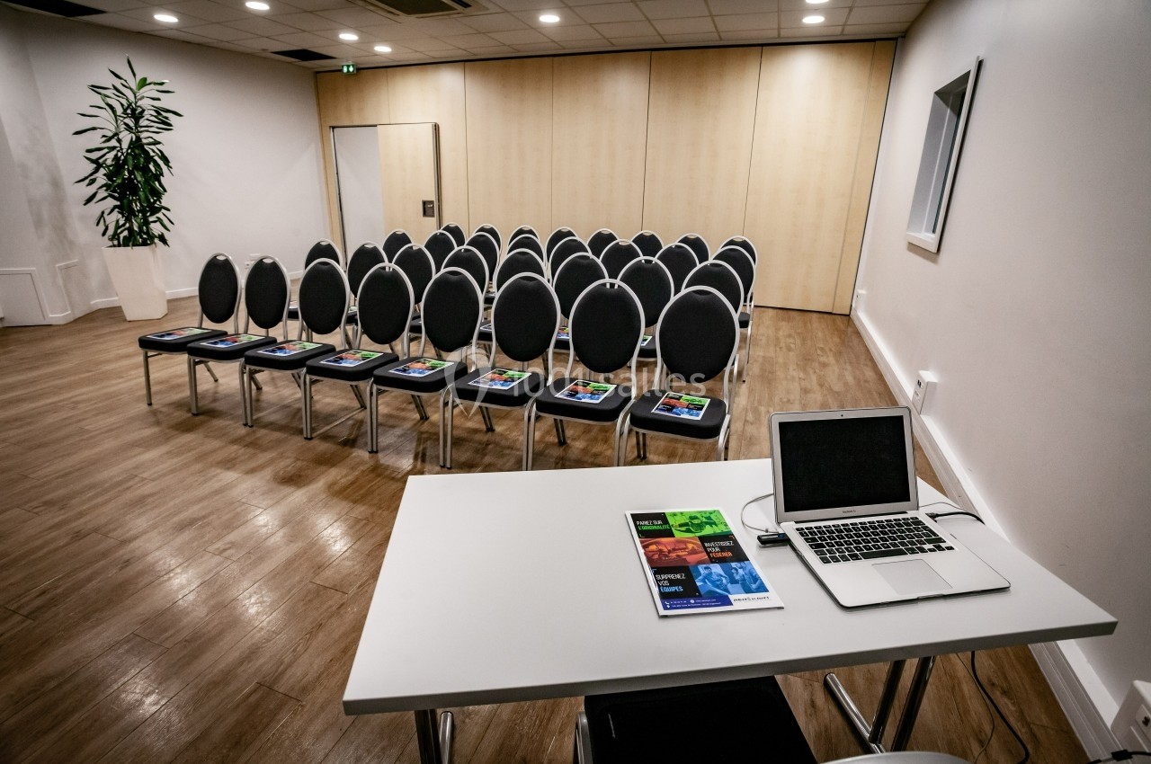 Salle de réunion avec chaises alignées, brochures posées sur chaque siège et un bureau équipé d'un ordinateur portable.