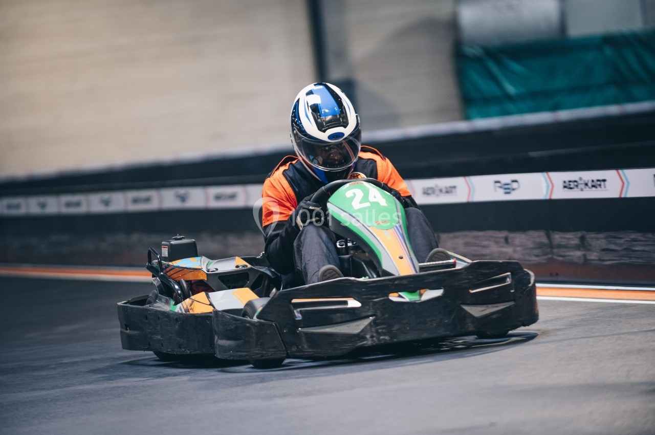 Un pilote en combinaison orange conduit un kart sur une piste intérieure.