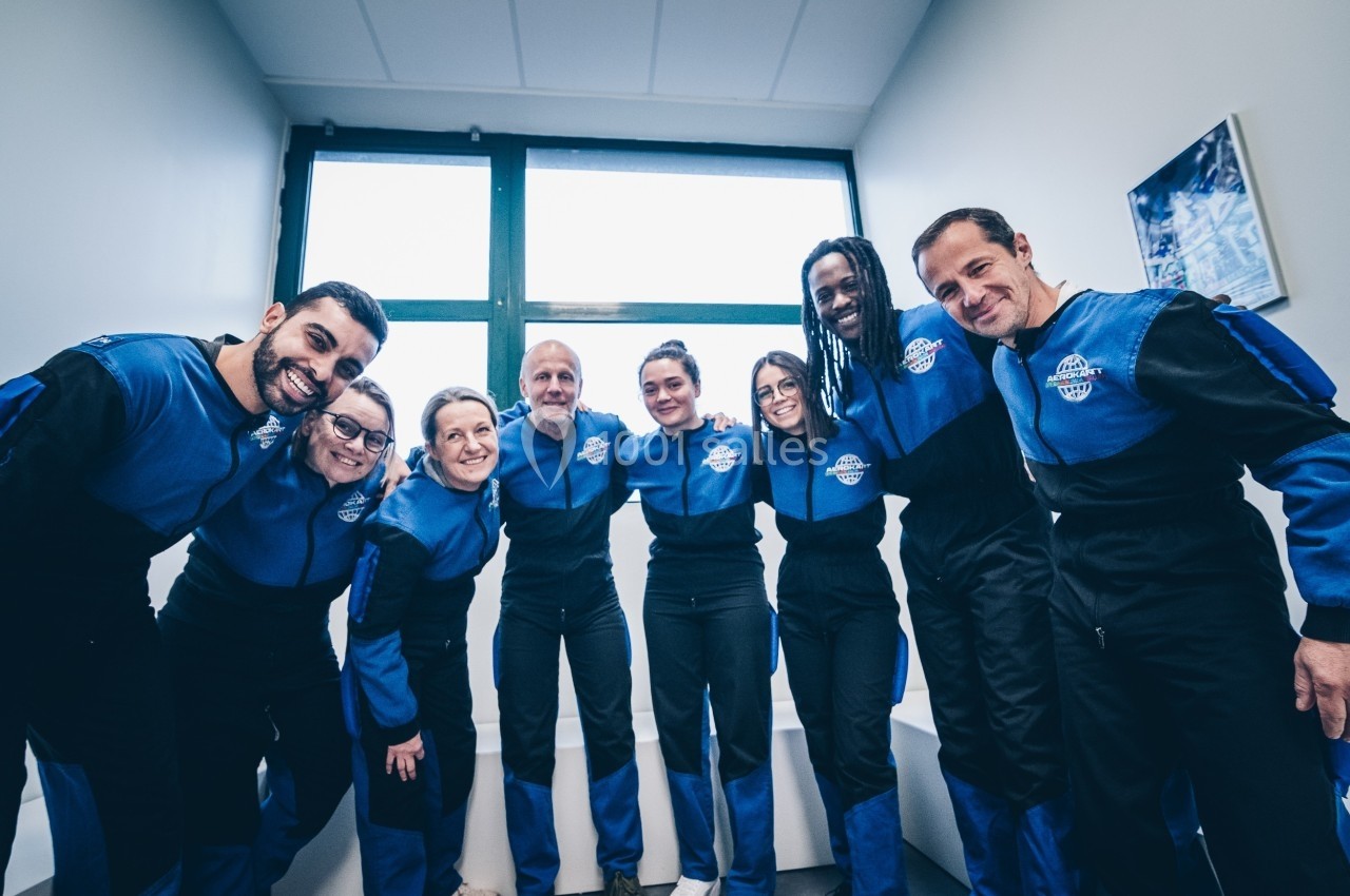 Un groupe de personnes en combinaison bleue sourit et pose dans une pièce lumineuse avec une grande fenêtre.