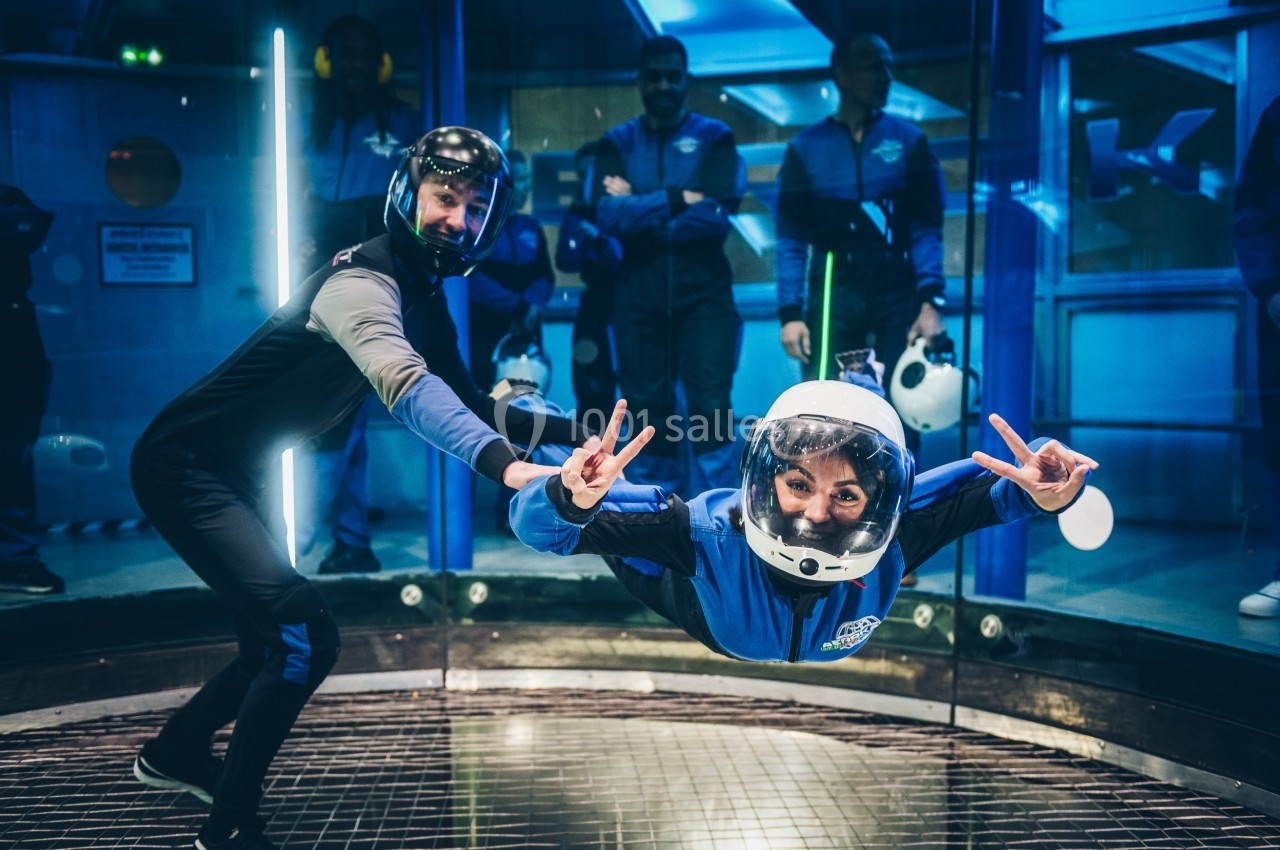 Une femme en combinaison bleue flotte dans un simulateur de chute libre, guidée par un instructeur.