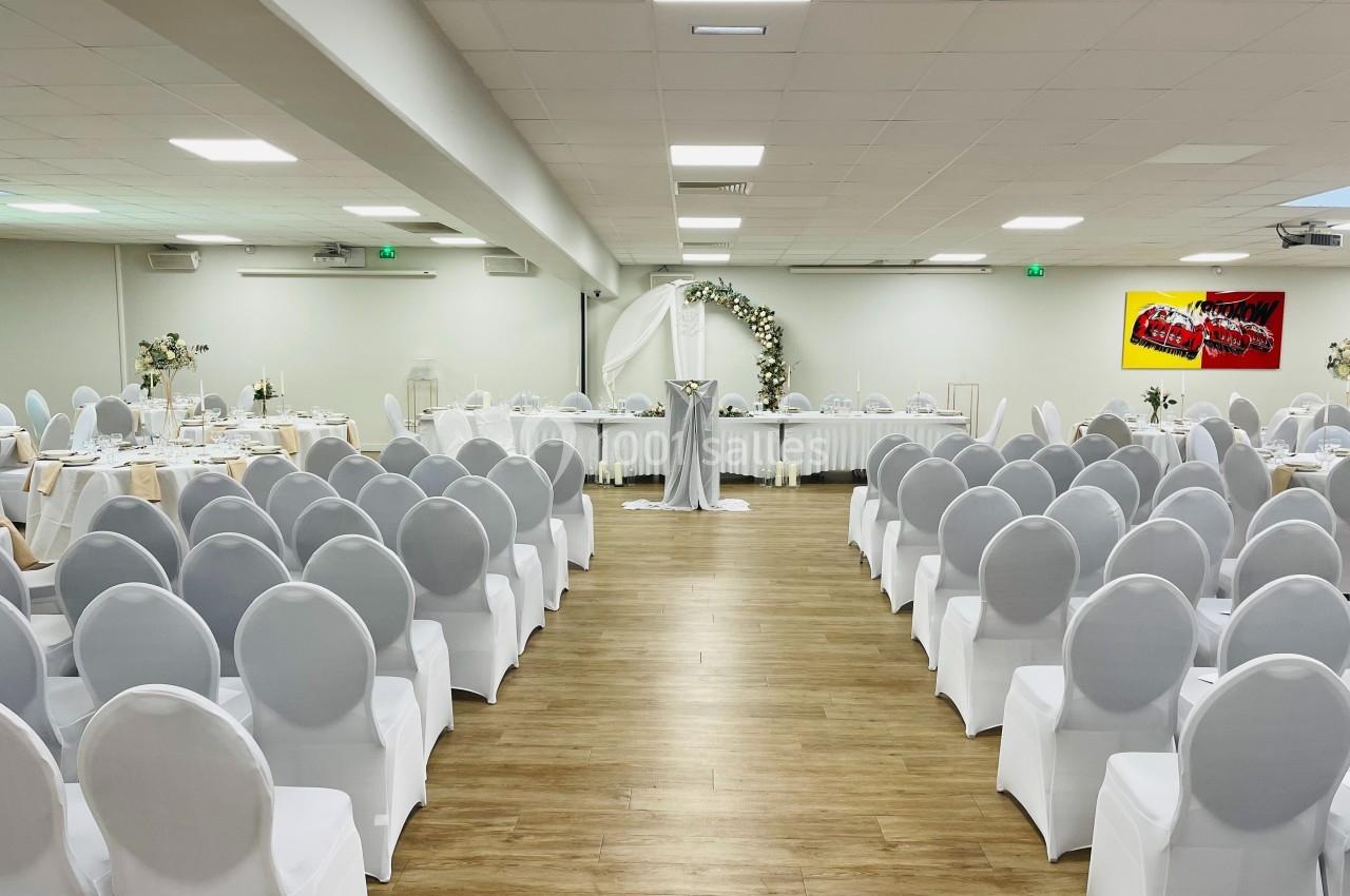 Salle de réception décorée pour un événement, avec des chaises blanches alignées et une arche florale au fond.