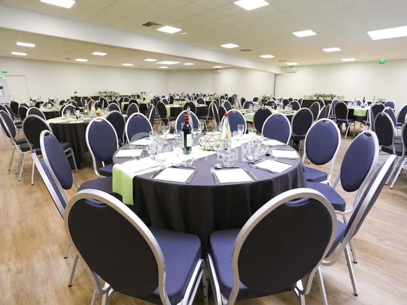 Salle de réception avec tables rondes dressées, nappes noires, chaises bleues et bouteilles de vin au centre.