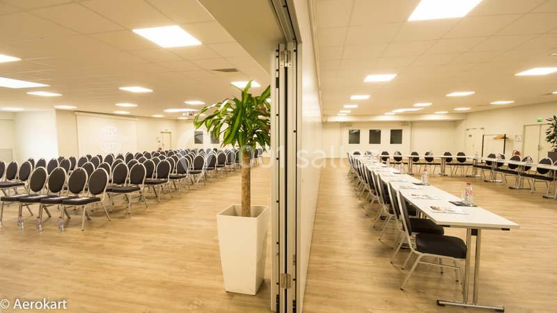 Salle de réunion lumineuse avec chaises alignées d'un côté et tables avec chaises de l'autre, séparées par une cloison…