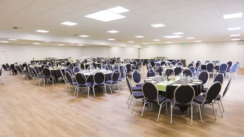 Salle de réception lumineuse avec tables rondes dressées, nappes vertes et chaises noires sur un sol en parquet.