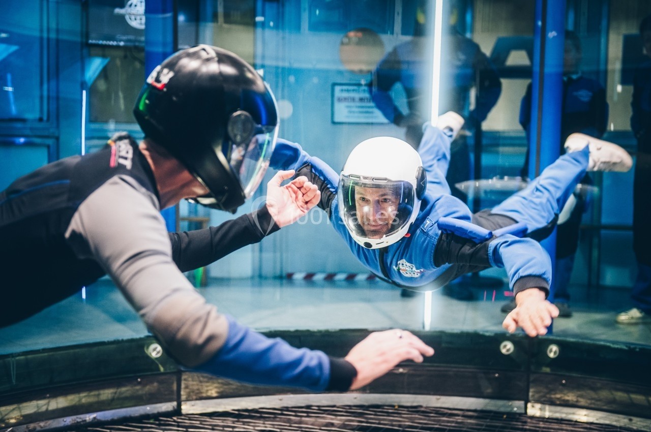 Un homme en combinaison bleue flotte dans un tunnel de vent, guidé par un instructeur en combinaison noire.