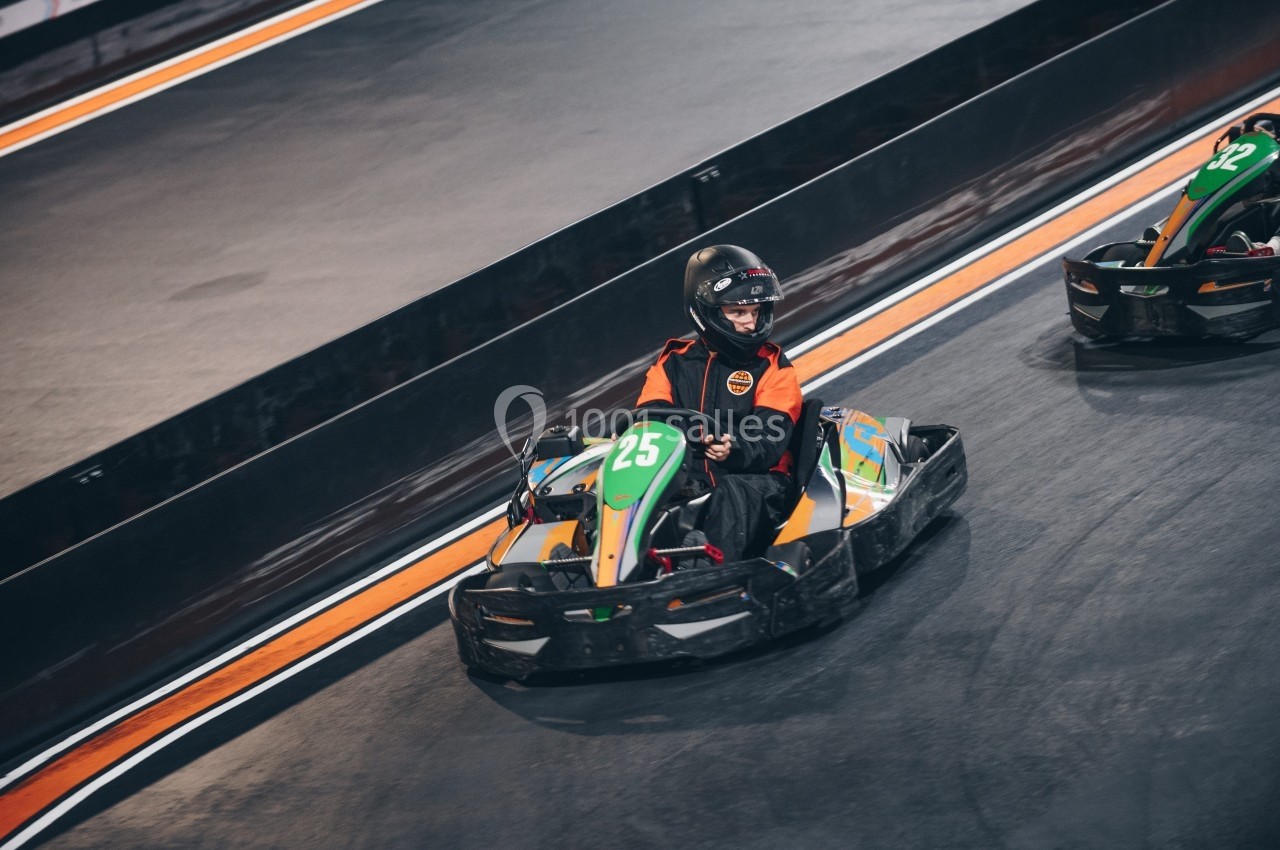 Une personne en karting portant un casque et une combinaison orange sur une piste intérieure.