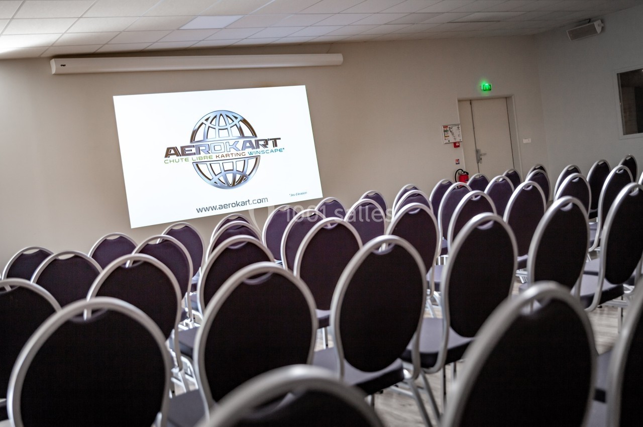 Salle de conférence vide avec des rangées de chaises noires et un écran affichant le logo ’Aerokart’.