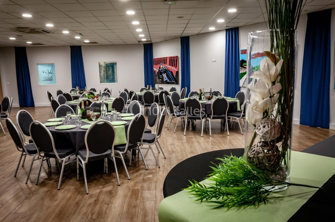 Salle de réception avec tables rondes dressées, décorée de nappes vertes et d'un grand vase avec des fleurs blanches.