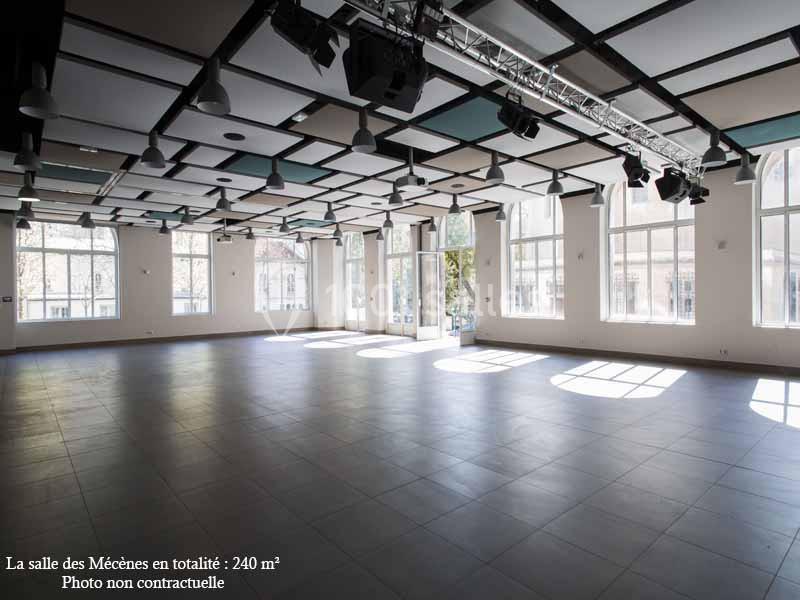 Salle spacieuse et lumineuse avec de grandes fenêtres, un plafond géométrique et un sol en carrelage sombre.