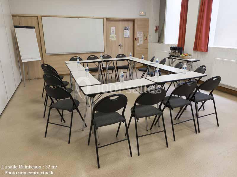 Salle de réunion avec tables disposées en U, chaises noires, tableau blanc et coin avec boissons et collations.