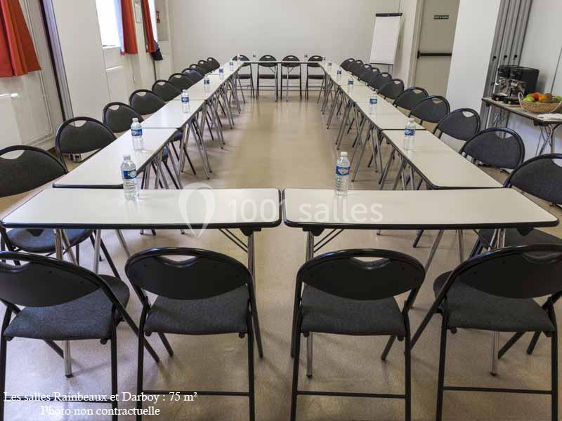 Salle de réunion en configuration en U avec des tables blanches, des chaises noires et des bouteilles d'eau disposées.