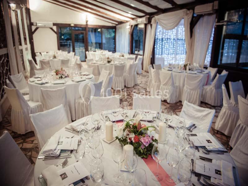 Salle de réception élégante avec tables rondes dressées, nappes blanches, décor floral et lumière naturelle.