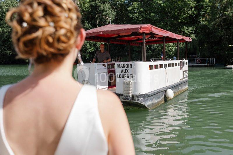 Une femme en robe blanche regarde un petit bateau nommé ’Manoir aux Loups II’ naviguant sur une rivière bordée d'arbres.