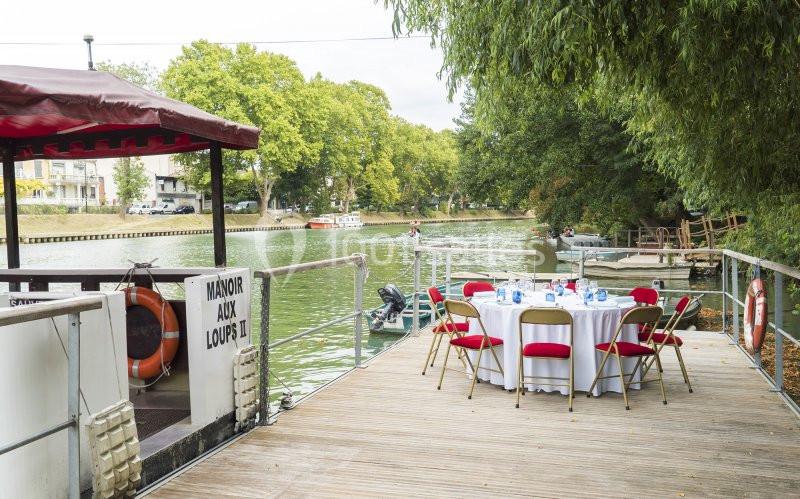 Table dressée sur un ponton en bois au bord d'une rivière, entourée de verdure et de bateaux amarrés.