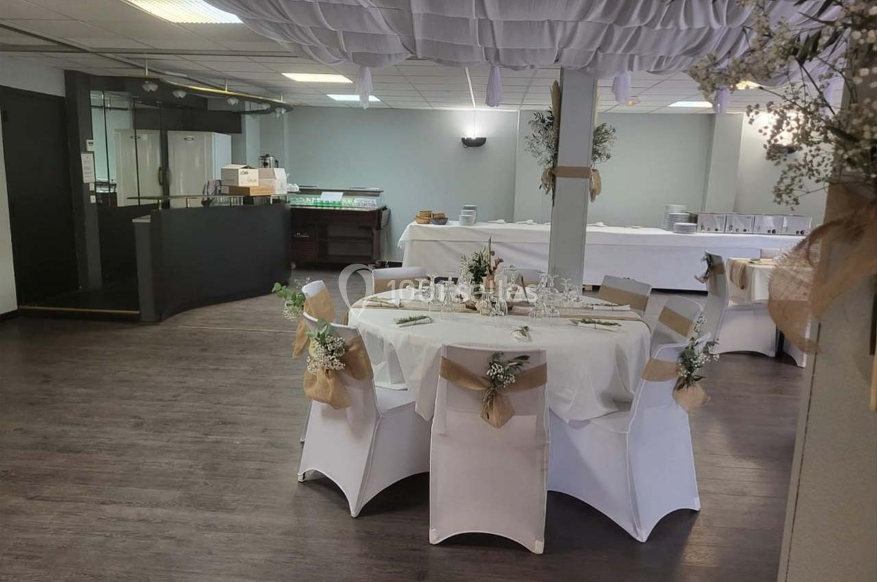 Salle de réception décorée avec des tables rondes, nappes blanches et chaises ornées de rubans et fleurs.