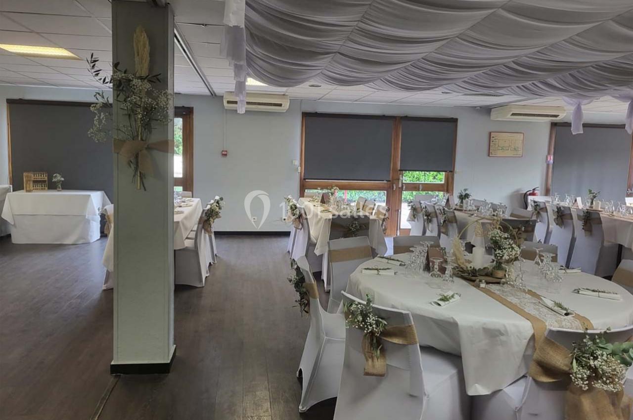Salle de réception décorée pour un événement, avec tables dressées, chaises blanches et ornements floraux.