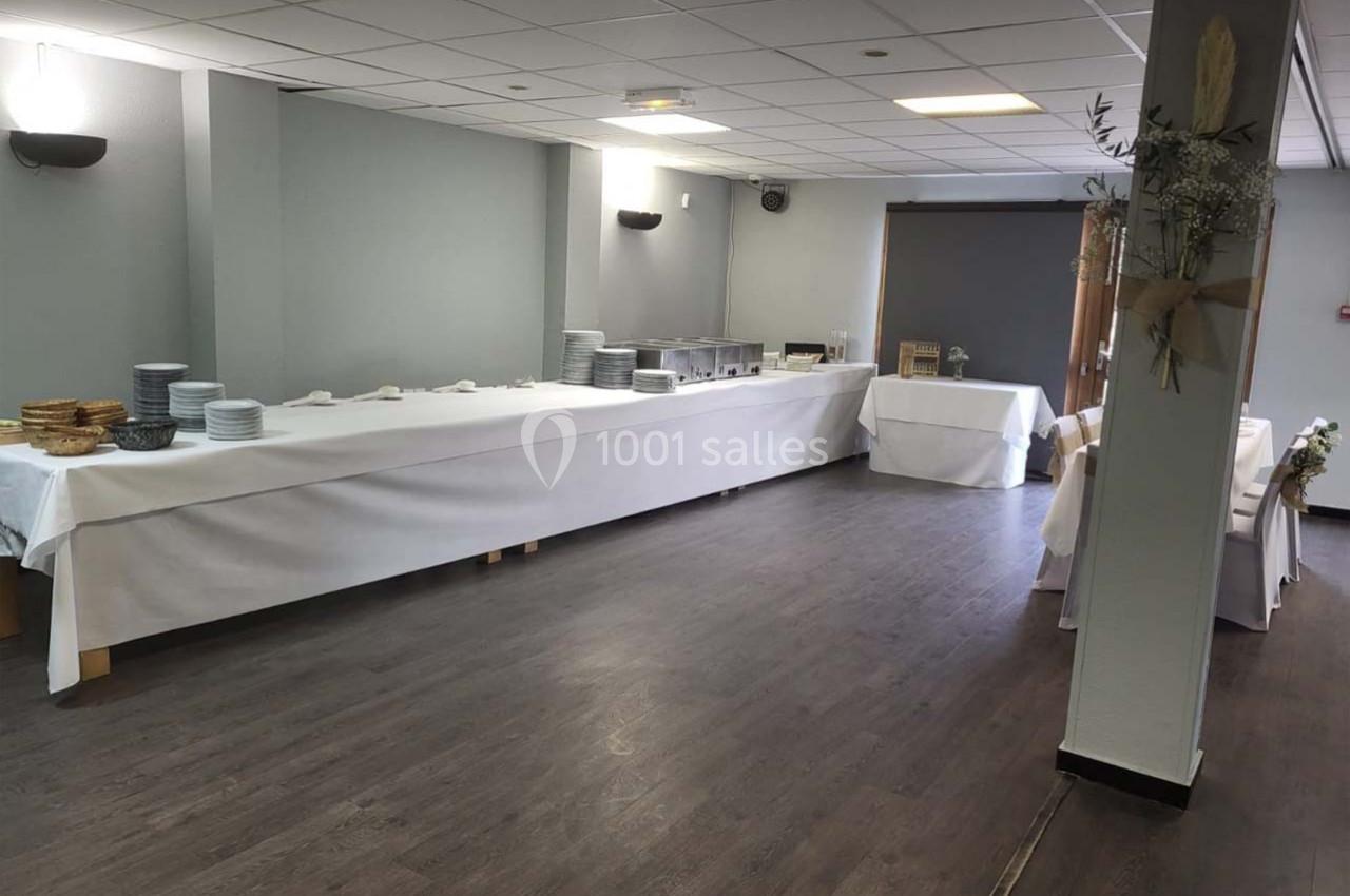 Salle avec tables alignées couvertes de nappes blanches, préparées pour un buffet avec assiettes et plats disposés.