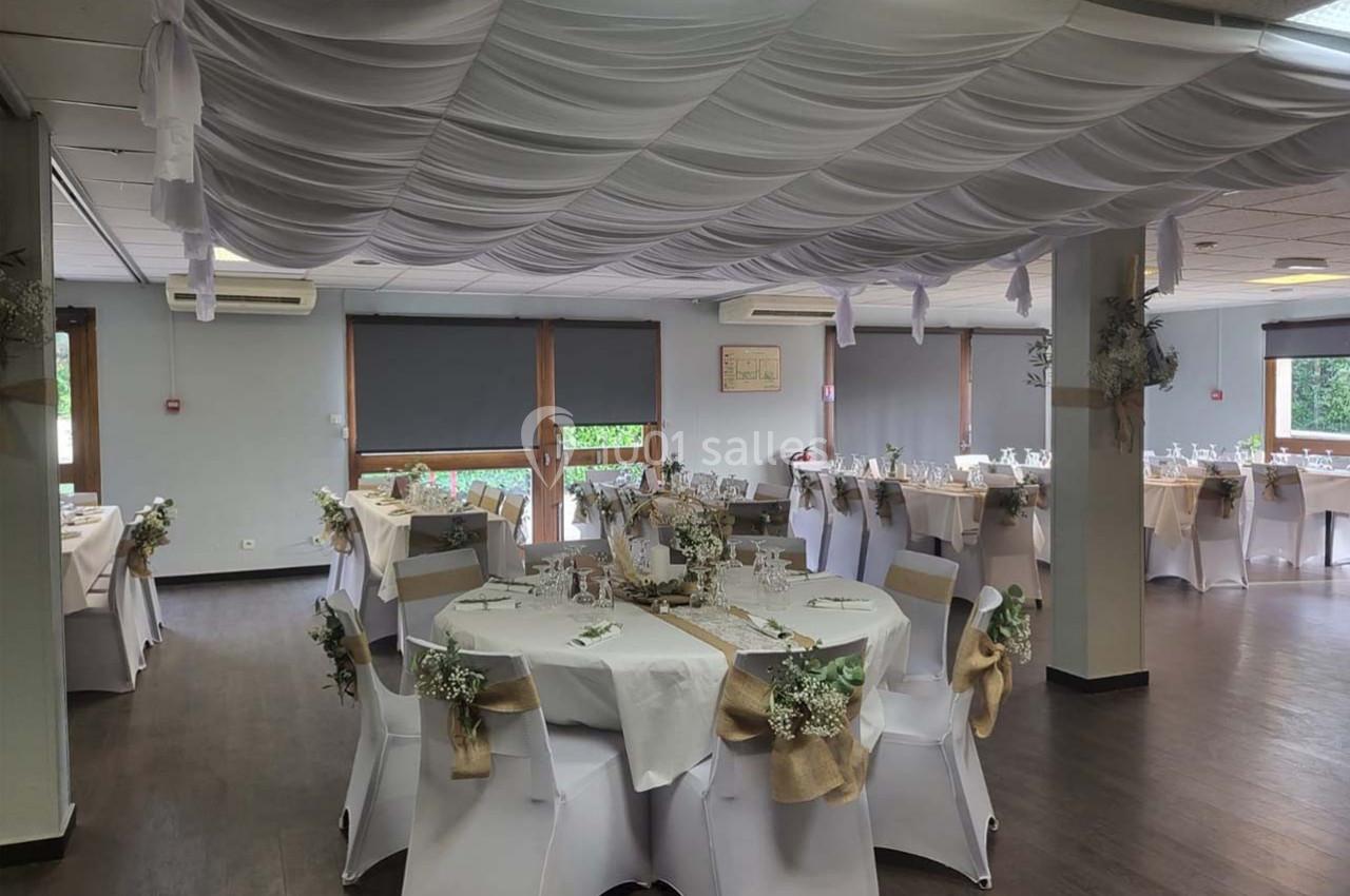 Salle de réception décorée pour un événement, avec tables rondes dressées, nappes blanches et ornements floraux.