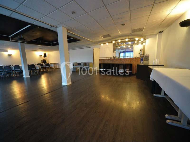 Salle spacieuse avec parquet sombre, bar éclairé au fond, tables et chaises disposées sur le côté.