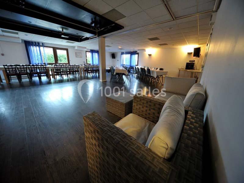 Salle spacieuse avec tables alignées, chaises, canapés en rotin au premier plan et grandes fenêtres en arrière-plan.