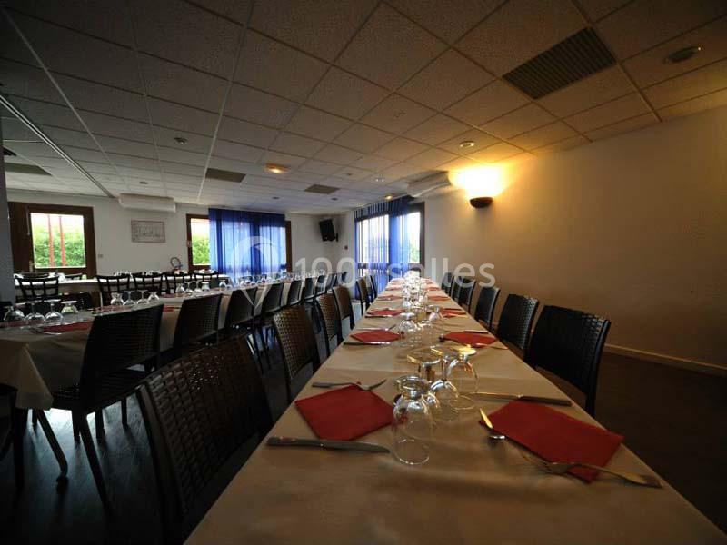 Salle de restaurant avec de longues tables dressées, nappes blanches, serviettes rouges et lumière naturelle entrant par les…