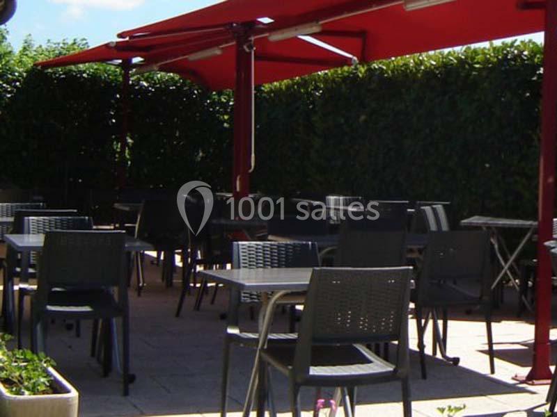 Terrasse extérieure avec tables et chaises en métal noir, ombragée par des auvents rouges et entourée de haies vertes.