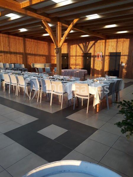 Salle de réception lumineuse avec de grandes tables dressées, chaises blanches et poutres en bois apparentes.