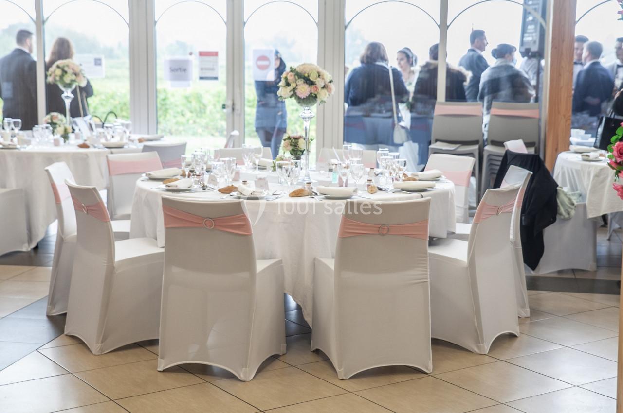 Salle de réception avec tables rondes dressées, nappes blanches, chaises décorées et invités en arrière-plan.