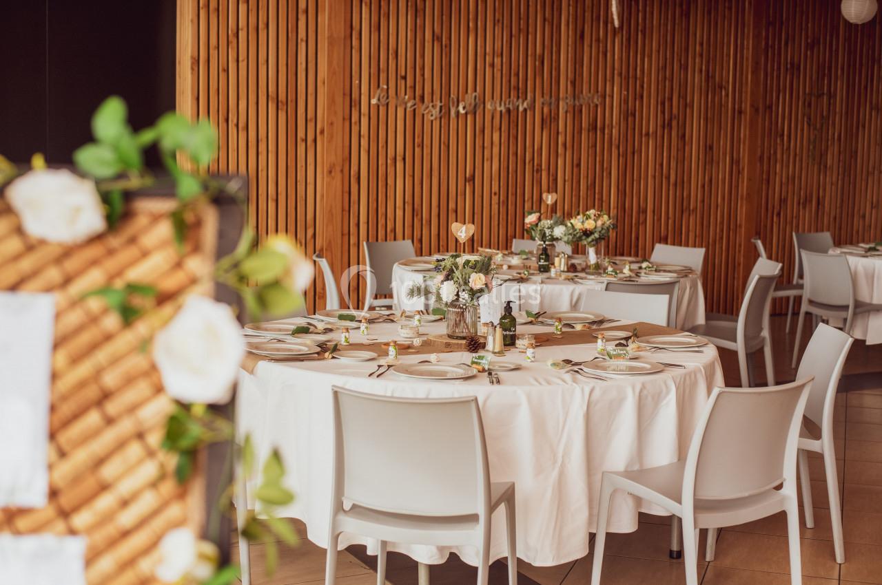 Salle décorée avec des tables rondes dressées, nappes blanches, fleurs et éléments de décoration élégants.